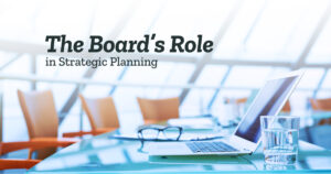 The Board's Role in Strategic Planning. Image shows an empty boardroom table, chairs, and an open laptop.
