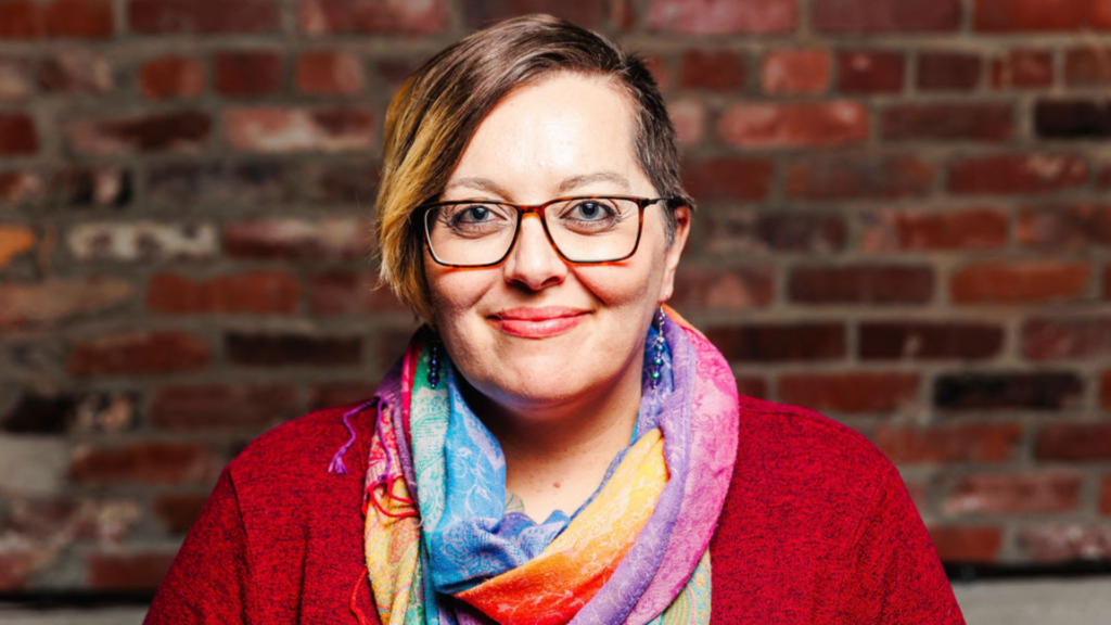 Taylor Boulware, PhD. Taylor is smiling wearing glasses, a rainbow scarf, and red sweater.