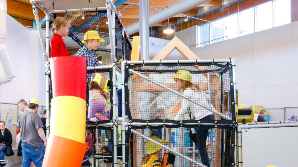 Scott Family Amazeum. Image shows children wearing play construction hats climbing on an indoor playground.