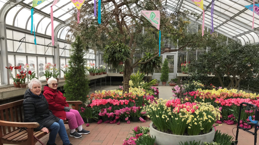 Evangelical Homes of Michigan. Image shows two women sitting on a bench in a colorful greenhouse.