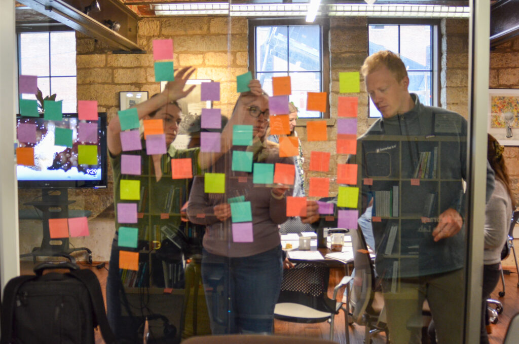 Image shows Benefactor Group team members reorganizing colorful post-it notes through a glass window.